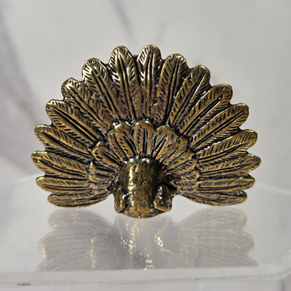 A small metallic decorative object shaped like a fan or open shell, with an antiqued gold/bronze finish, displayed on a clear acrylic stand against a light gray marble-like background.
