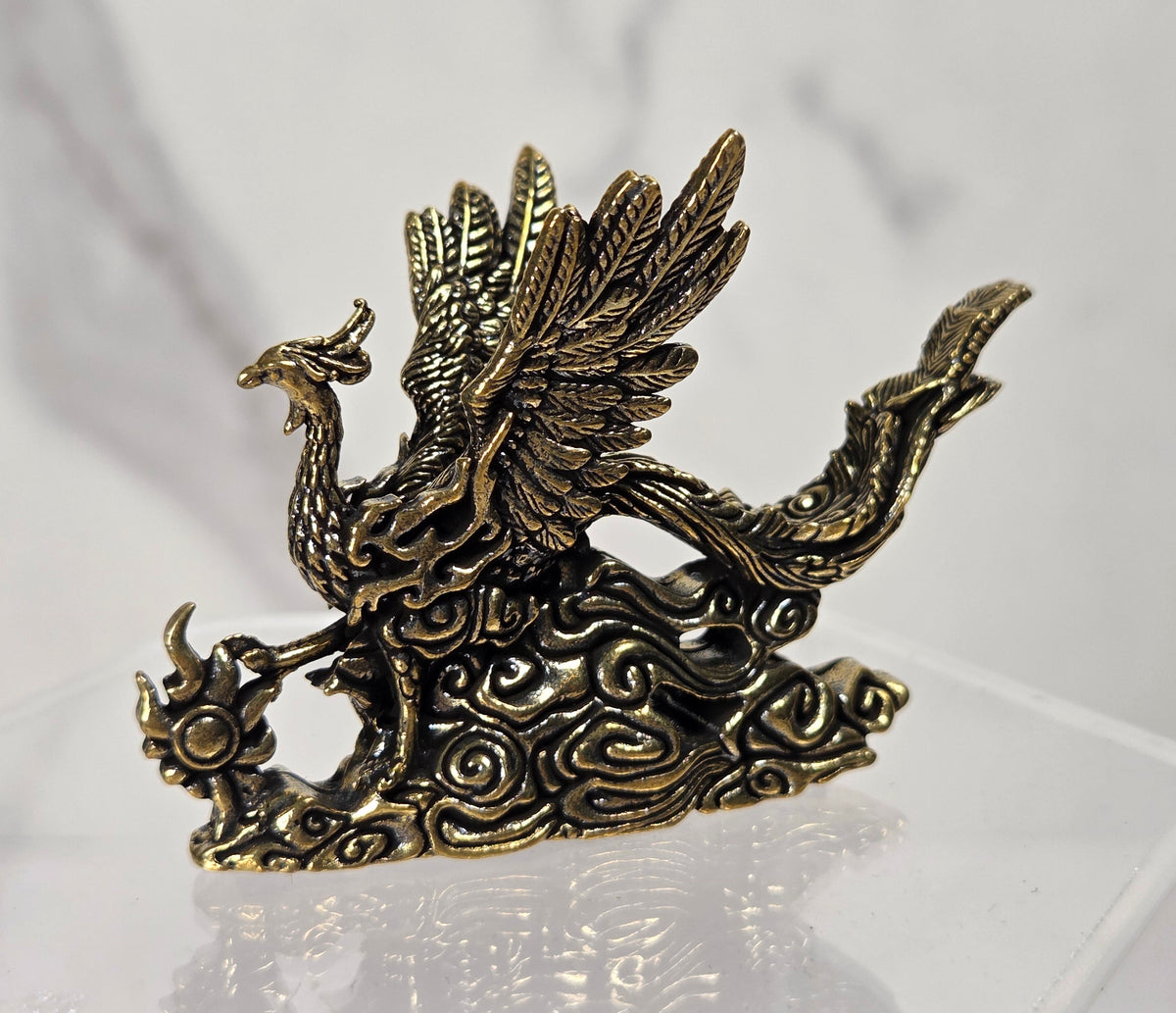 An antique bronze phoenix figurine standing on stylized clouds, displayed on a clear acrylic stand against a light gray marble-patterned background.