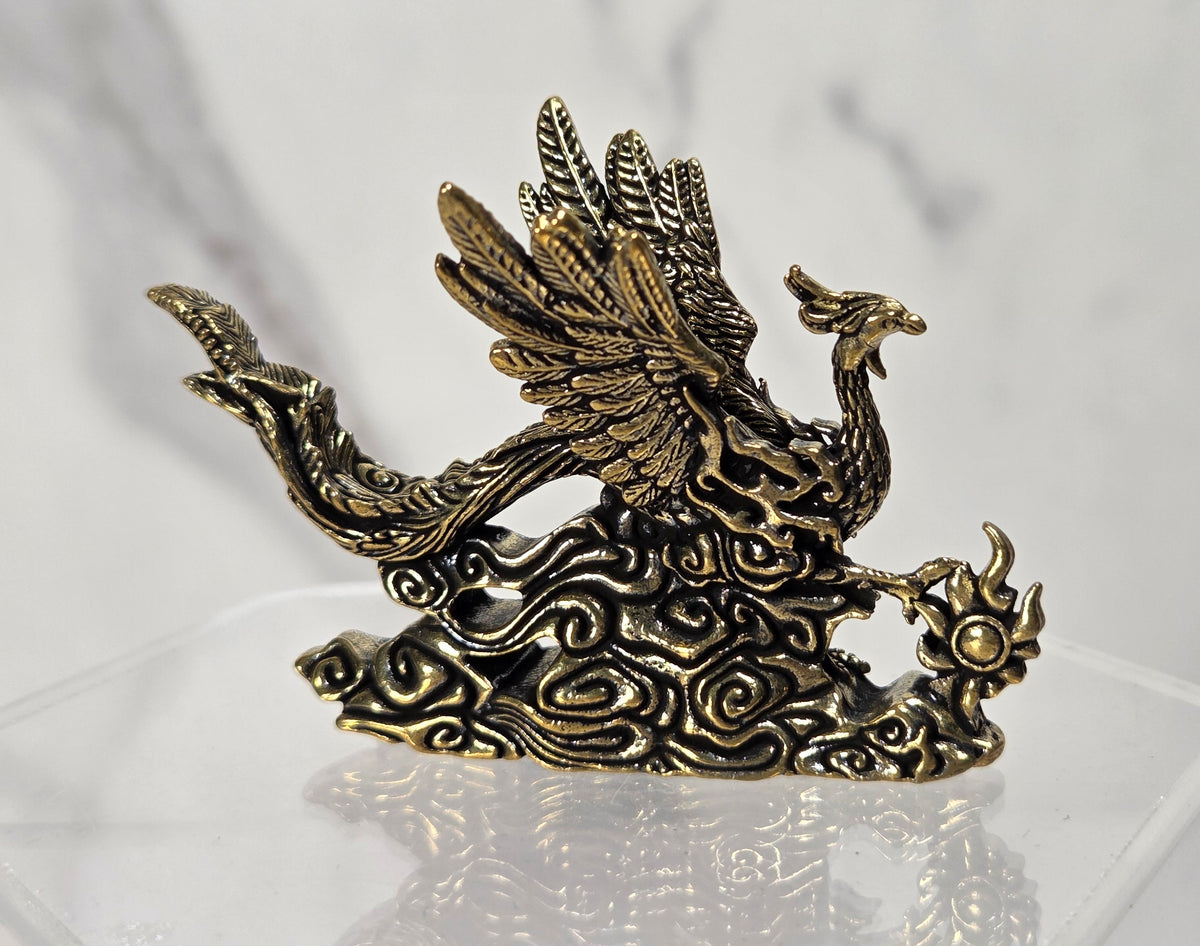 An ornate bronze-colored phoenix figurine with detailed feather and cloud motifs, displayed on a clear acrylic stand against a light gray marble-patterned background.