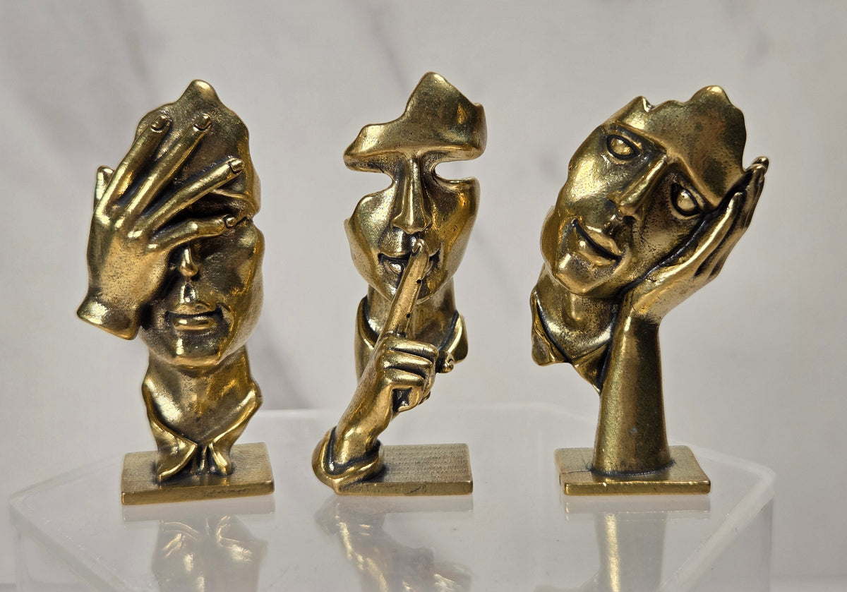 Three small metallic figurines shaped like stylized human faces and hands, displayed on a clear acrylic stand against a light gray marble-patterned background.