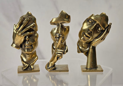 Three small metallic figurines shaped like stylized human faces and hands, displayed on a clear acrylic stand against a light gray marble-patterned background.