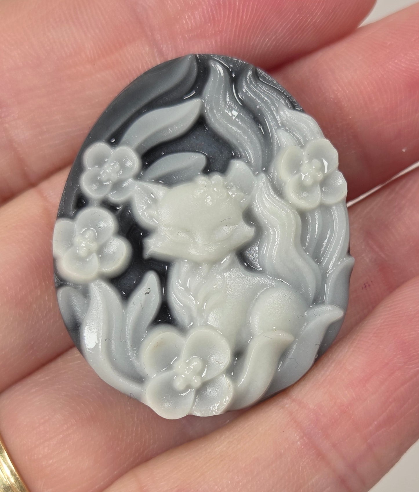 A hand holds an oval carved stone pendant featuring a fox surrounded by flowers, with a dark gray and white marbled pattern.