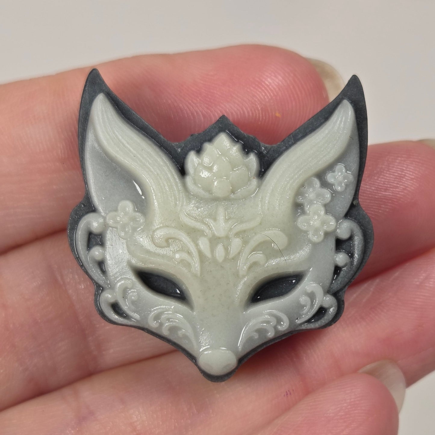 A small carved fox-head pendant or charm with floral details, held in a person's hand. The item appears off-white/ivory with dark gray to black outlining and shadows.