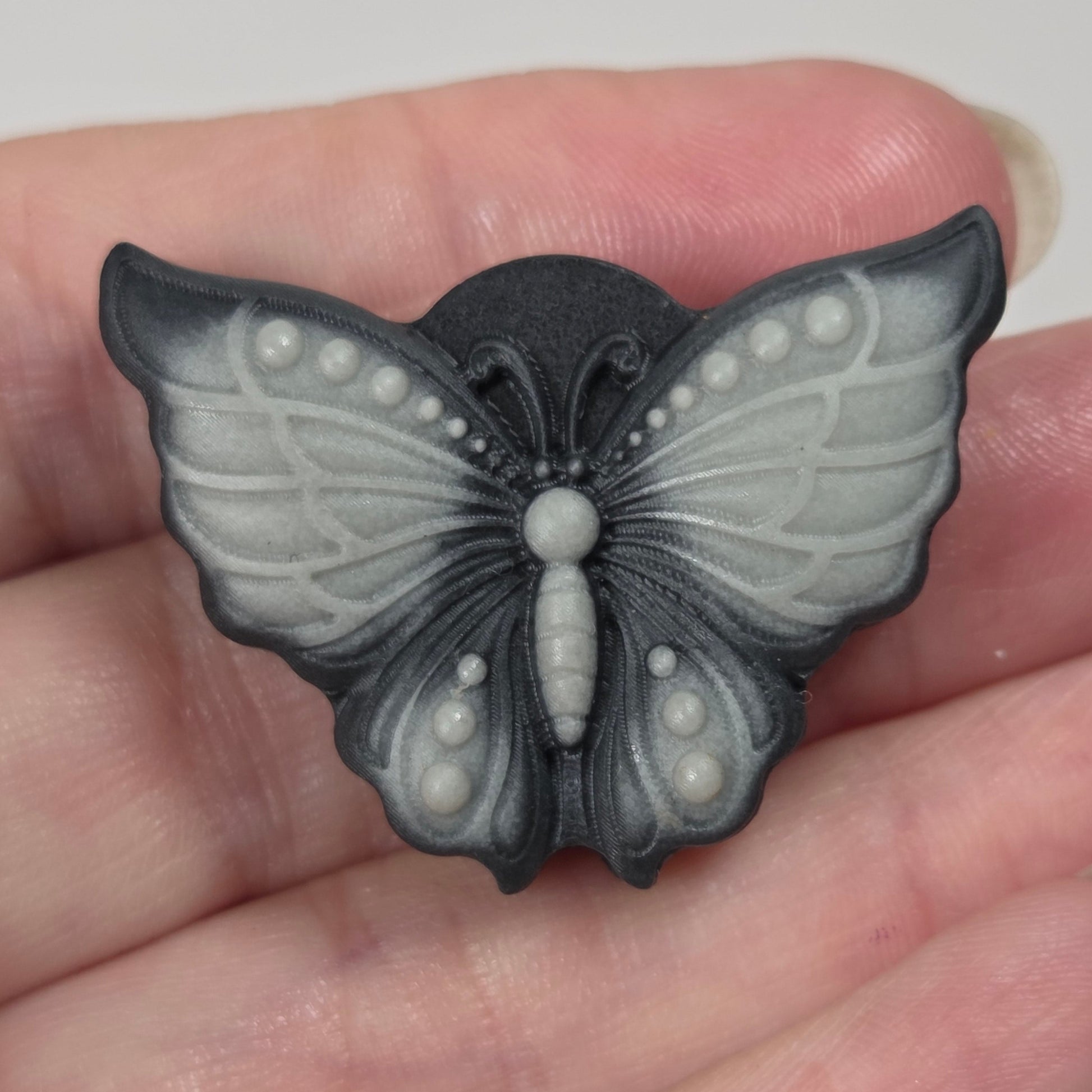 A butterfly-shaped jewelry piece with a dark charcoal gray and light gray marbled pattern, held in a person's hand.