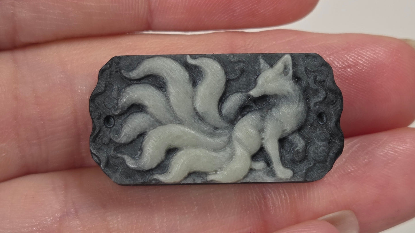 A small carved rectangular pendant or charm featuring a fox with flowing tails, held between fingers. The pendant is made of a dark gray stone or resin with a lighter gray raised fox design, shown against a soft off-white background.