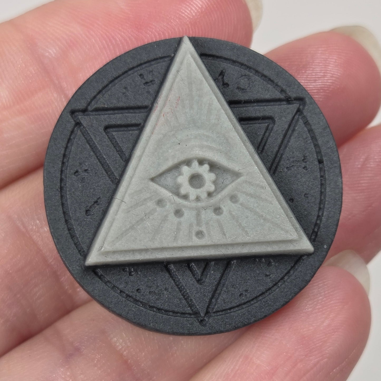 A circular dark gray medallion featuring a raised light gray triangle with an eye motif in the center, held in a person's hand against a plain light background.