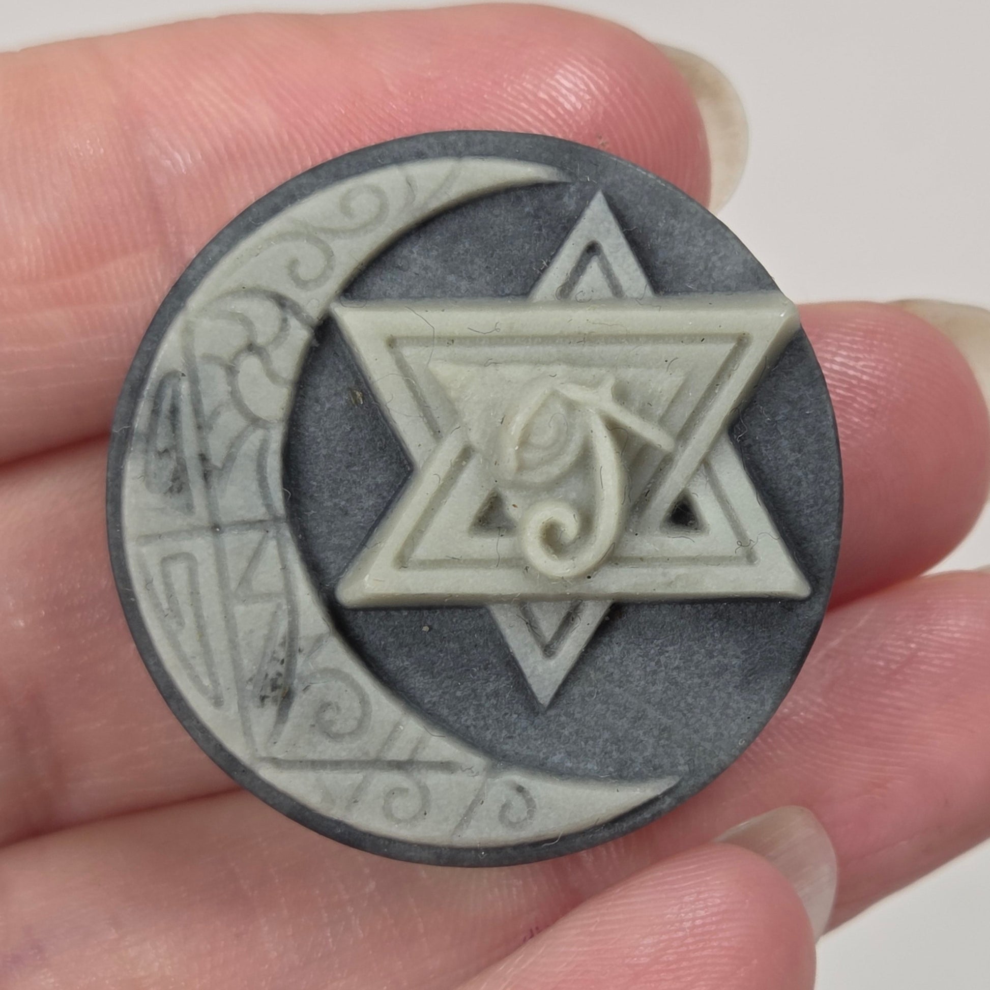 A hand holding a circular dark gray and off-white medallion featuring a crescent moon, a Star of David, and a stylized Hebrew letter Shin.