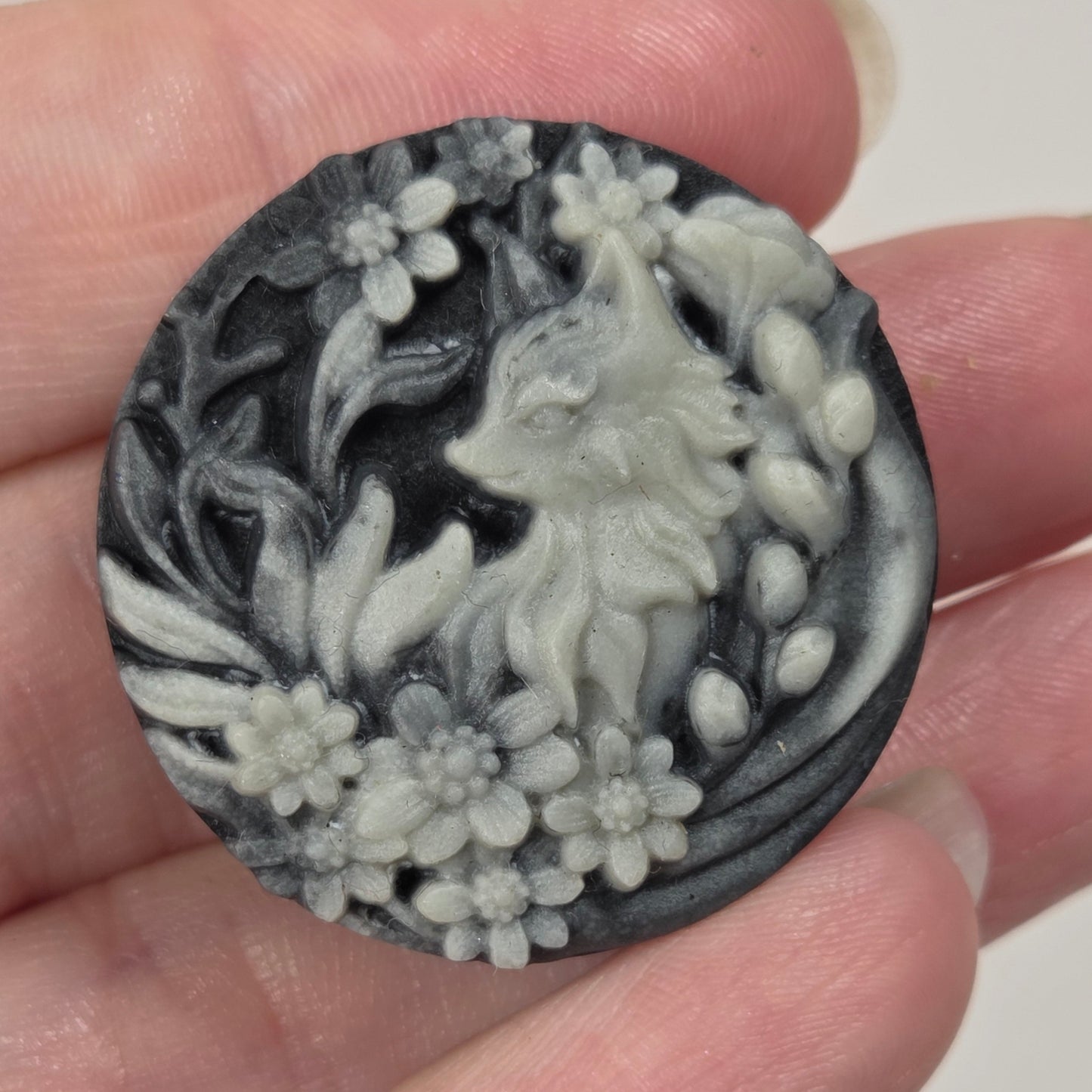 A round carved stone or resin cabochon featuring a fox surrounded by flowers, held in a person's hand. The cabochon has a dark charcoal and light gray marbled pattern.