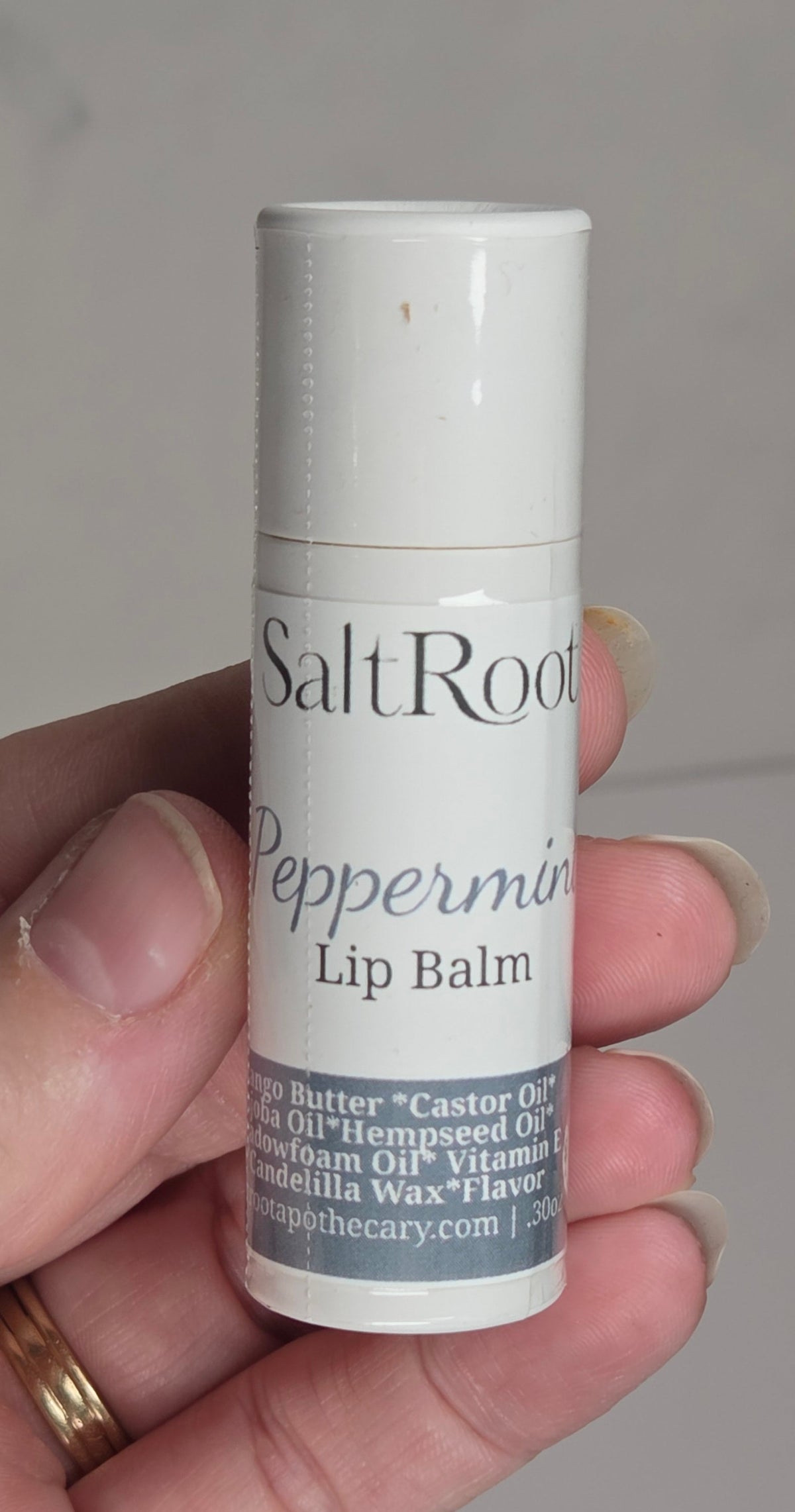 A hand holding a small white tube of Salt Root Peppermint Lip Balm against a soft gray background.