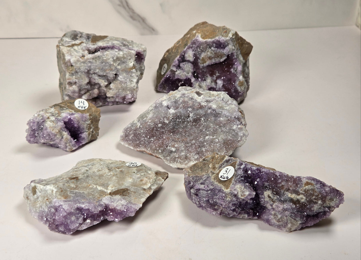Six raw amethyst geode specimens of various sizes arranged on a white surface with a light gray marble-patterned background. Several pieces show deep purple crystal cavities and others display rough, textured amethyst outer layers with tan matrix.