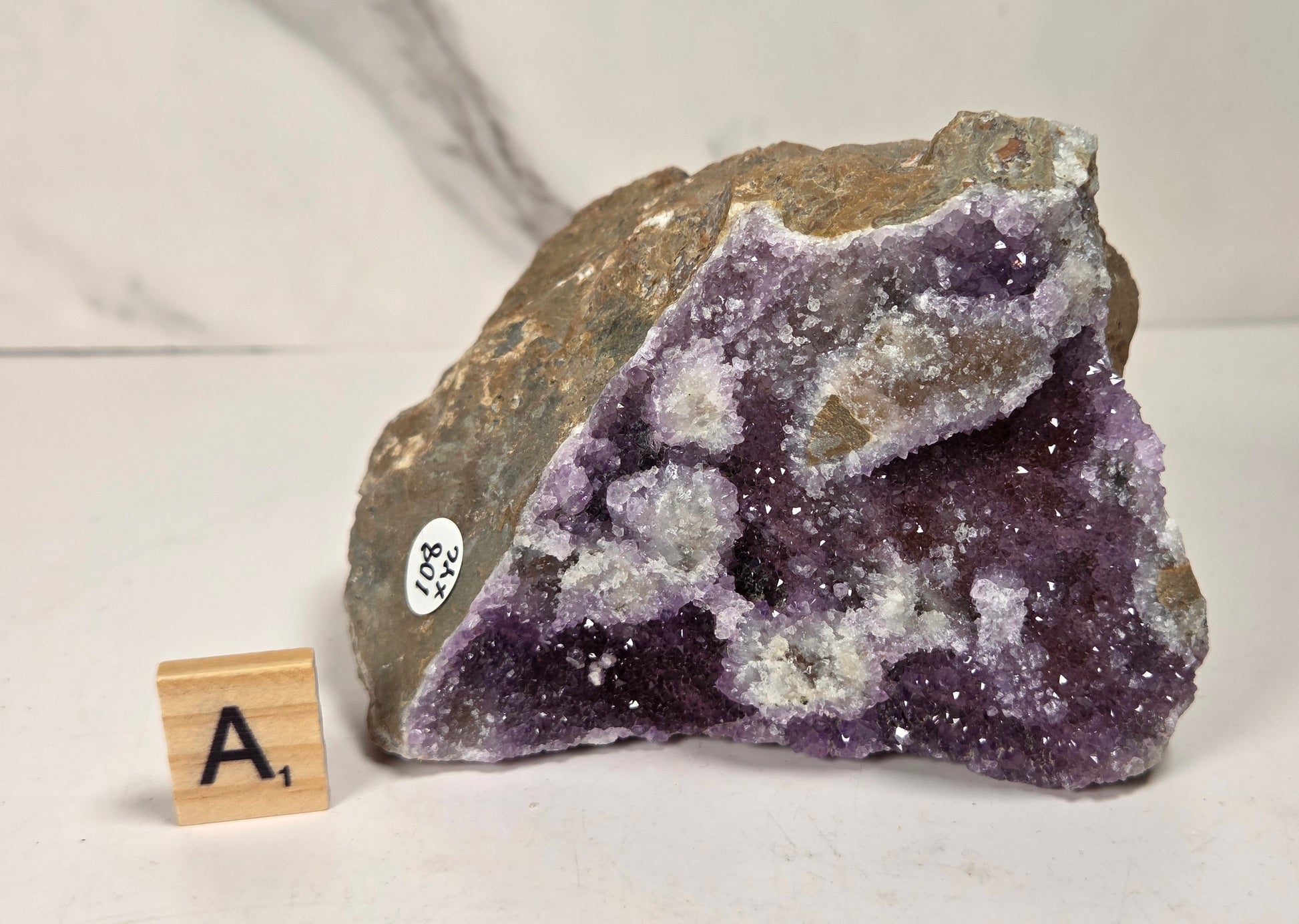 A raw amethyst geode specimen with a triangular section showing deep purple crystal clusters and white quartz points, set against a light gray marble-patterned background. A small wooden letter tile with the letter 'A' is placed next to it for scale.