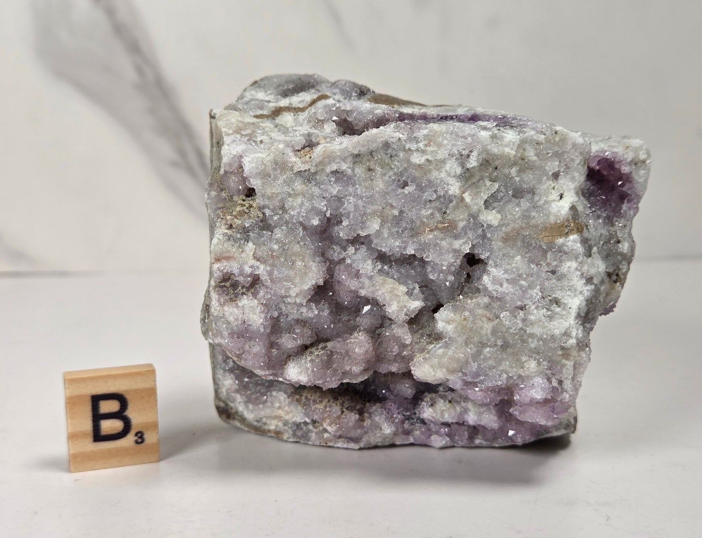 A rough crystal specimen with pale lavender and white crystalline structure on a gray matrix, shown next to a small wooden Scrabble tile marked B's on a light surface.