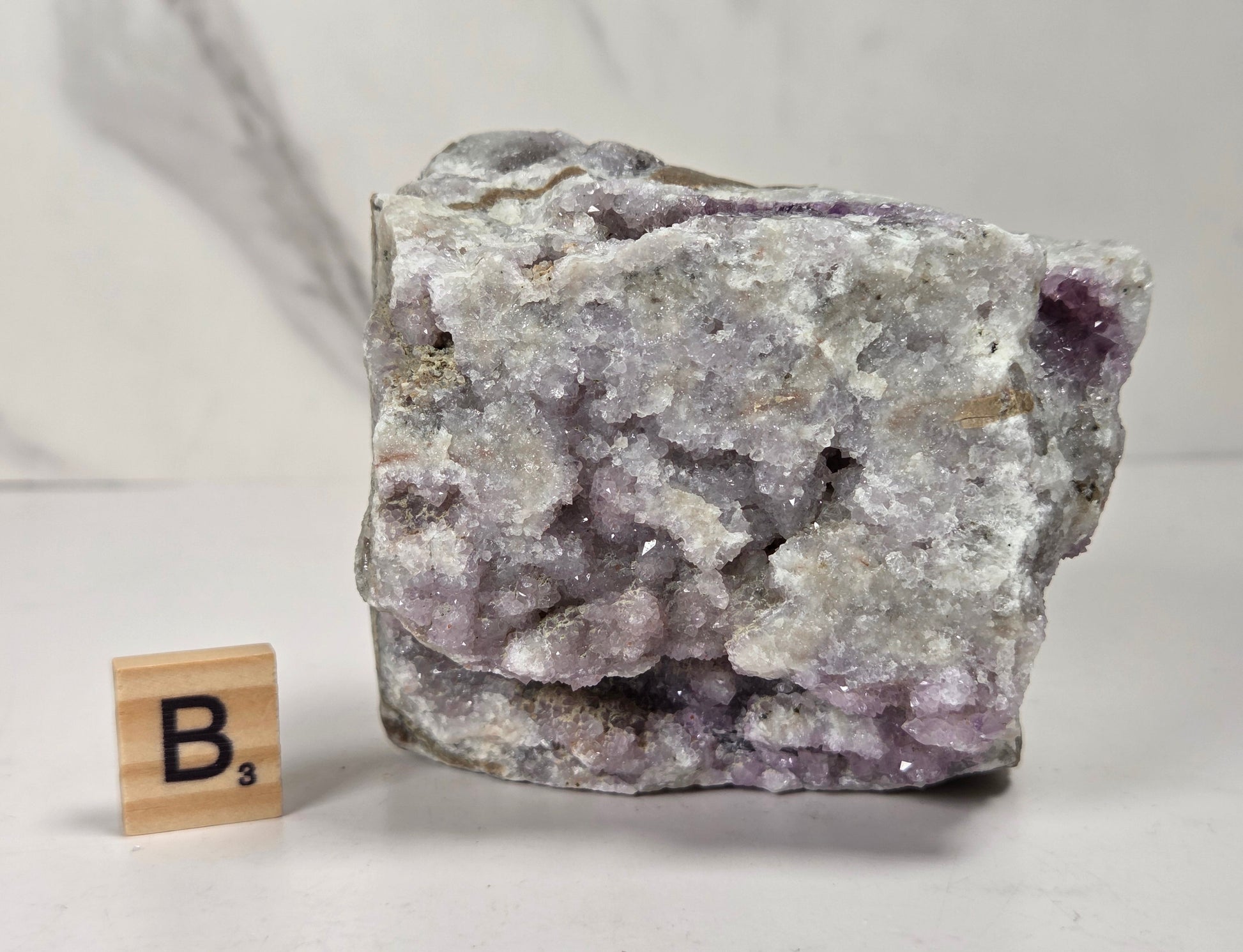 A rough crystal specimen with pale lavender and white crystalline structure on a gray matrix, shown next to a small wooden Scrabble tile marked B's on a light surface.