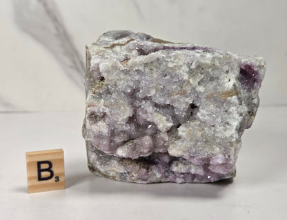 A rough crystal specimen with pale lavender and white crystalline structure on a gray matrix, shown next to a small wooden Scrabble tile marked B's on a light surface.
