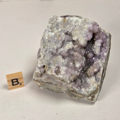 A raw amethyst geode specimen with light purple crystal clusters and a gray matrix, shown on a light surface next to a small wooden letter tile.