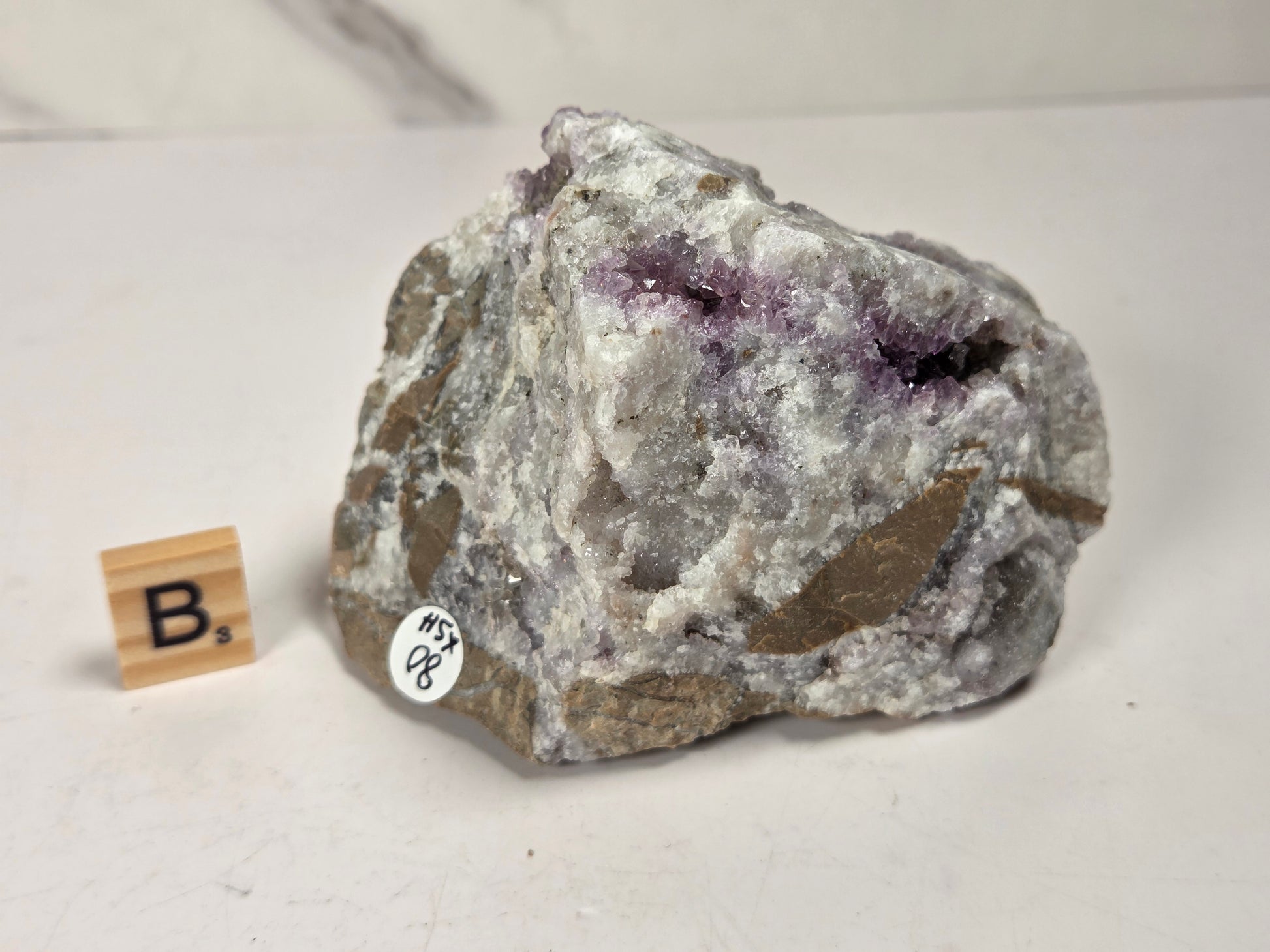 A raw amethyst geode specimen with purple crystal clusters on a gray and white matrix, displayed on a light surface next to a small wooden letter block and a white inventory sticker.