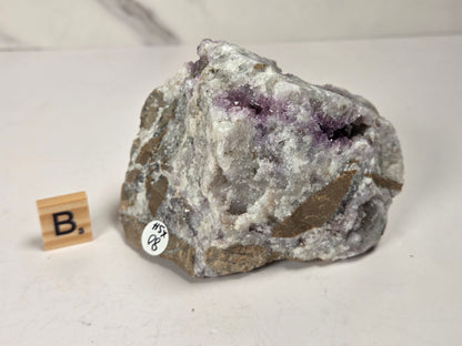 A raw amethyst geode specimen with purple crystal clusters on a gray and white matrix, displayed on a light surface next to a small wooden letter block and a white inventory sticker.