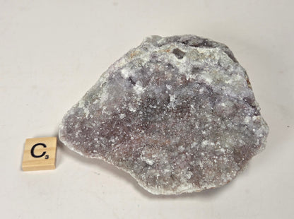 A rough, irregular crystal formation or mineral specimen with a white crystalline surface and gray-brown interior, photographed on a plain light background. A small wooden tile marked "C's" sits beside it for scale.
