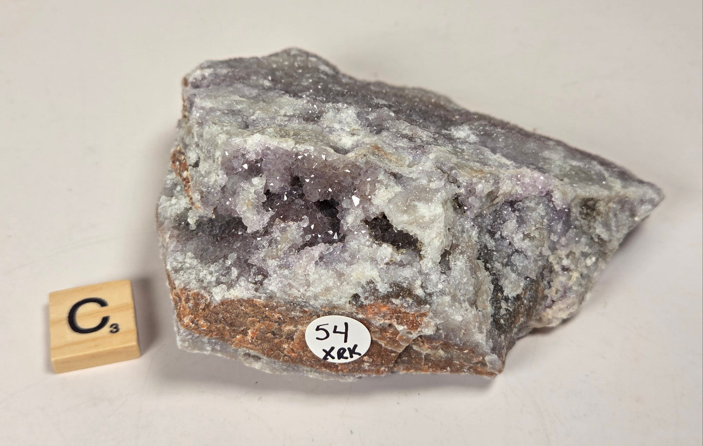 A raw mineral specimen with a purple crystal cavity surrounded by gray and white crystalline material and a brown matrix, shown on a light beige background next to a small wooden Scrabble tile and a circular sticker reading '54 XRX'.