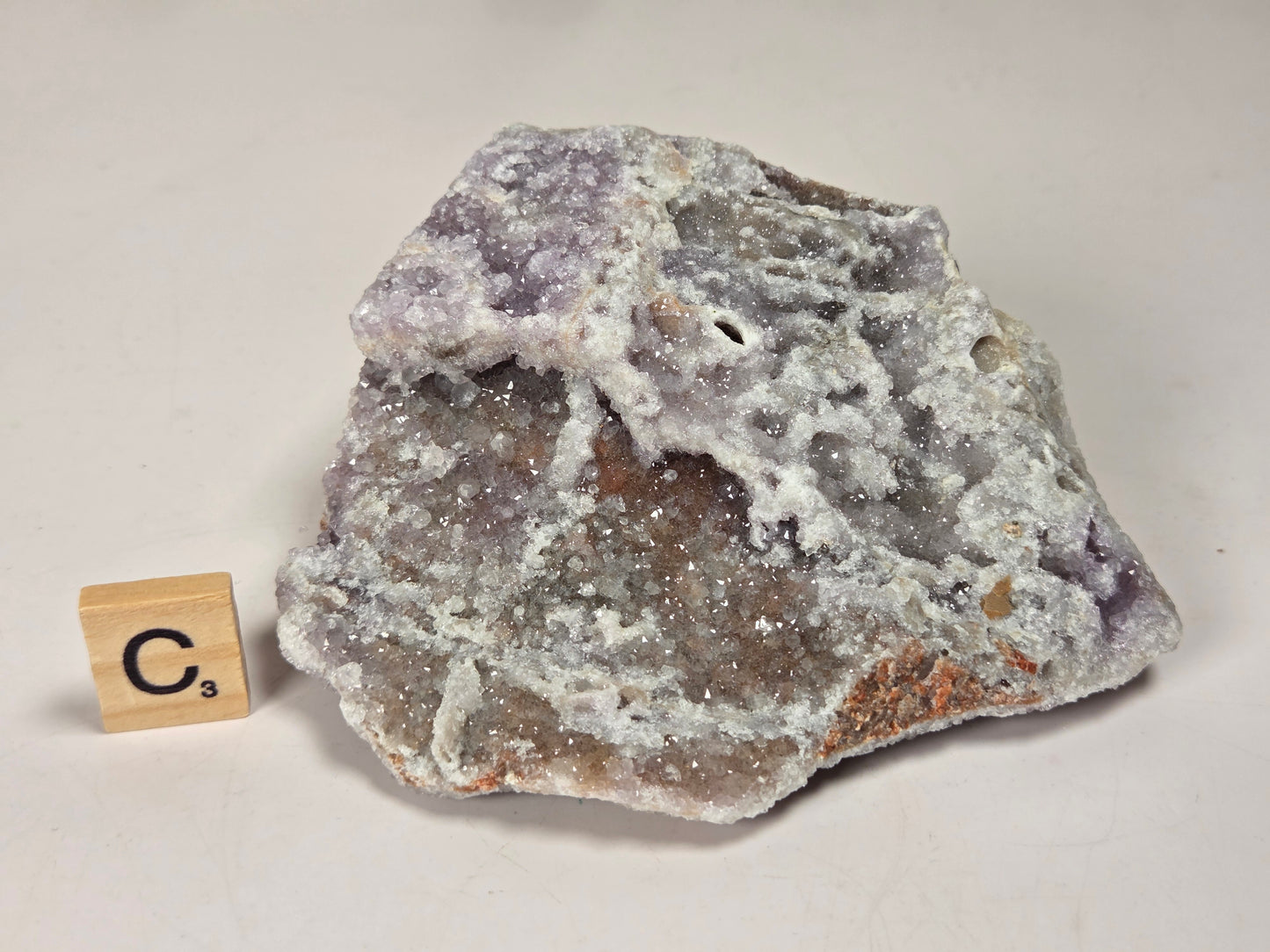 A rough natural mineral specimen with a white crystalline surface and purple-brown interior, photographed on a plain light background. A small wooden tile with the letter C is placed beside it for scale.