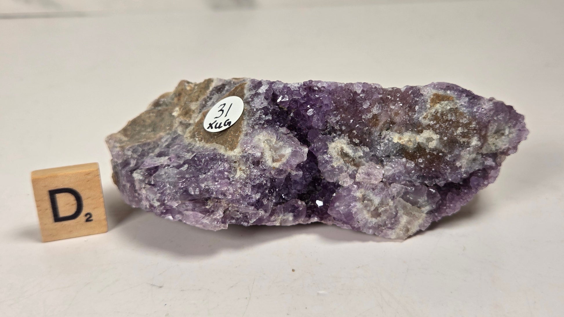 A raw purple amethyst geode specimen with white quartz inclusions and a rough brown matrix, displayed on a light surface next to a small wooden Scrabble tile showing the letter D and value 2.