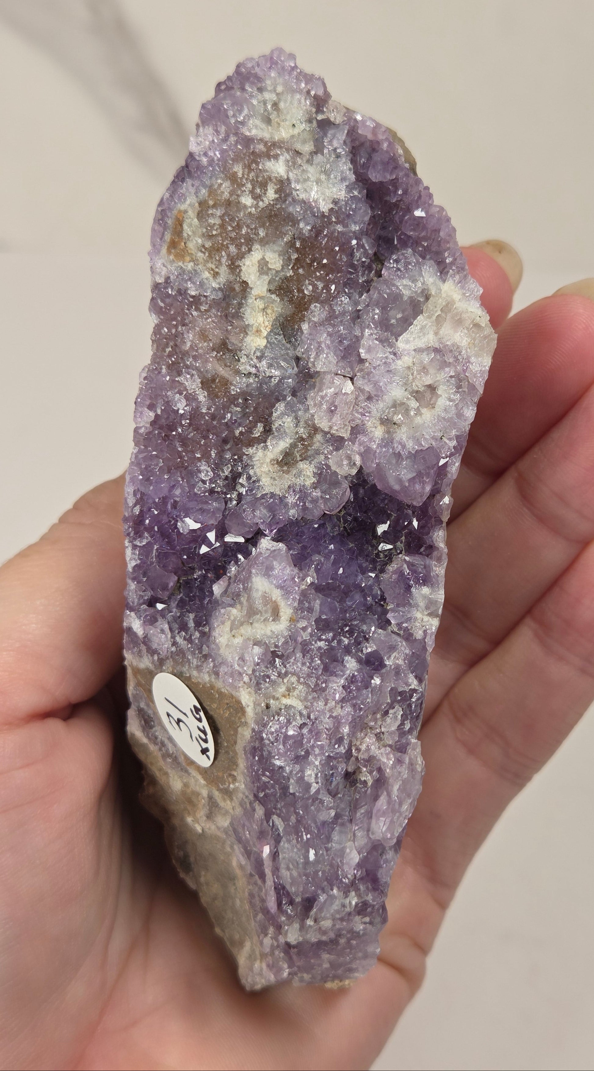 A hand holding a raw amethyst crystal cluster with a small identification label, set against a light marble-patterned background.