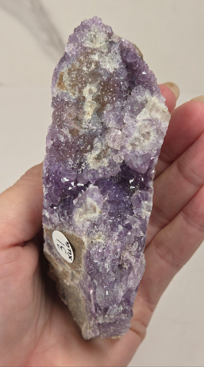 A hand holding a raw amethyst crystal cluster with a small identification label, set against a light marble-patterned background.