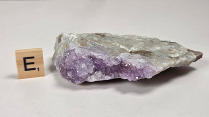 A raw amethyst crystal cluster with visible purple and white crystalline structure and a rough gray-brown outer matrix, photographed on a light neutral surface next to a small wooden letter tile showing the letter E.