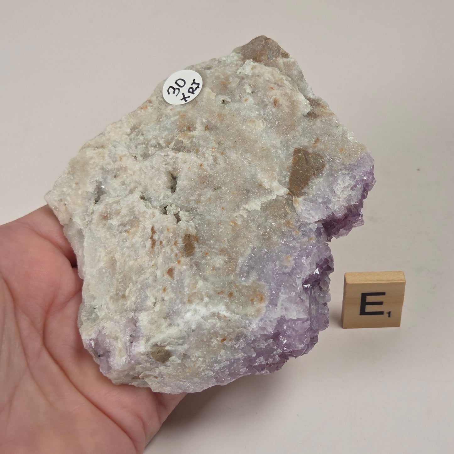 A hand holds a rough amethyst geode specimen showing a white and purple crystal structure, with a small wooden letter block labeled 'E1' beside it on a light surface.