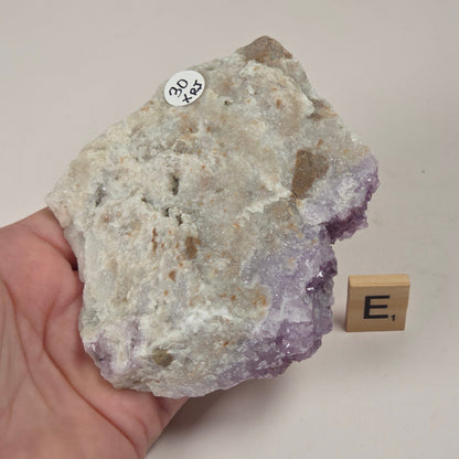A hand holds a rough amethyst geode specimen showing a white and purple crystal structure, with a small wooden letter block labeled 'E1' beside it on a light surface.