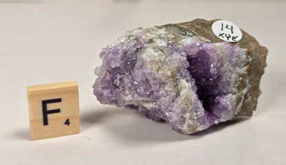 A raw amethyst geode specimen with a purple crystal cavity and rough outer matrix, displayed on a light surface next to a wooden letter tile showing 'F' and a small white label reading '14 XPE'.