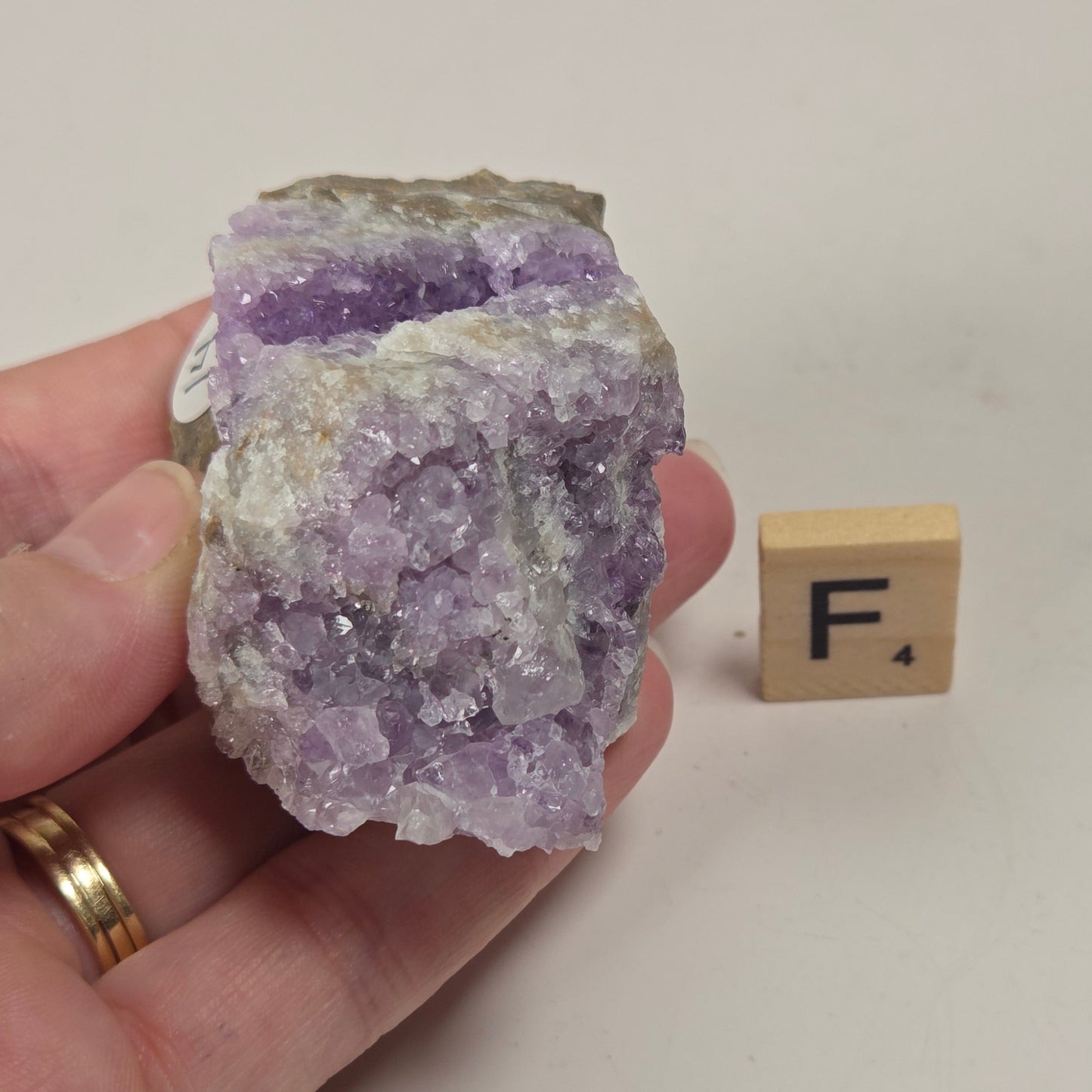 A hand holds a raw amethyst crystal cluster with visible purple crystal points and a gray matrix background, next to a small wooden letter block showing the letter F, on a plain off-white surface.