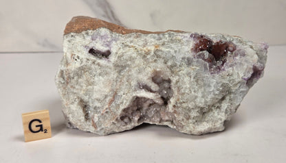 A raw mineral specimen with white and gray crystalline structure, brown matrix, and small purple inclusions, photographed on a light surface with a marble-look background. A small wooden tile with 'G2' is placed beside it for scale.