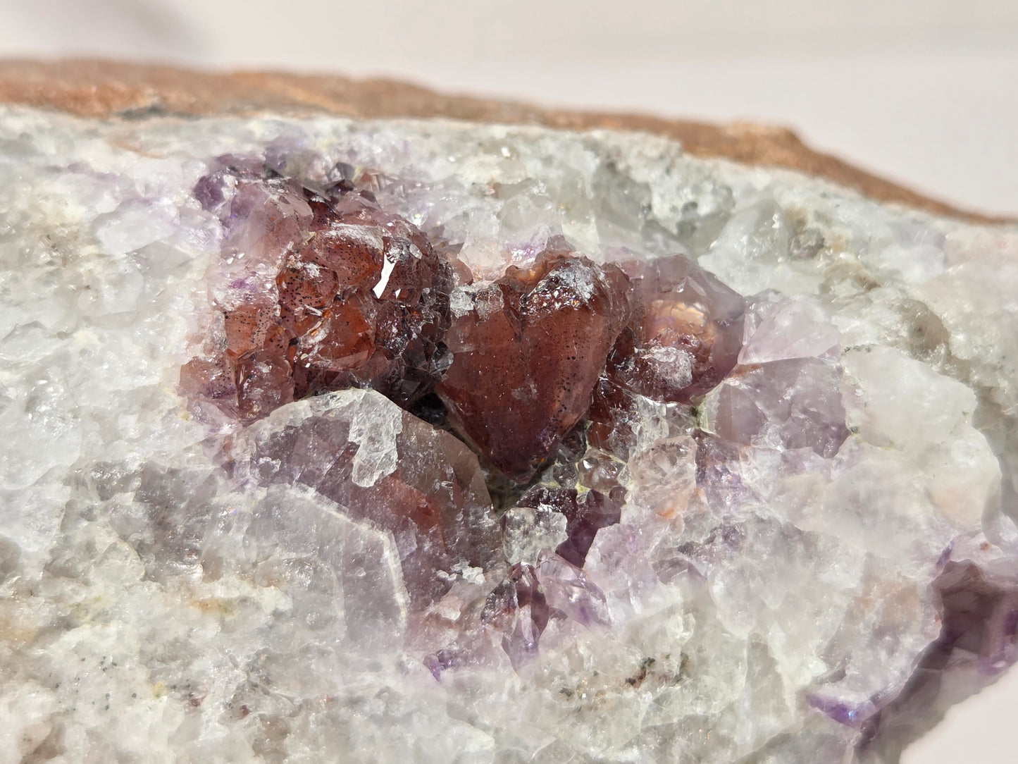 A close-up of a natural crystal cluster featuring translucent white and pale purple quartz with prominent reddish-brown amethyst crystal formations in the center, set against a neutral light background.