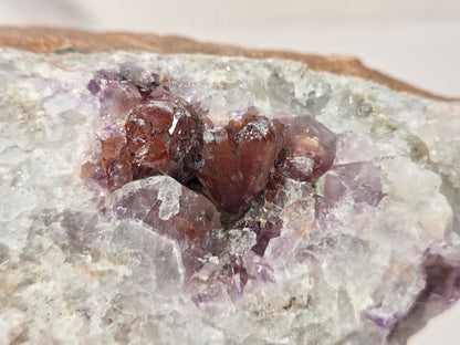A close-up of a natural crystal cluster featuring translucent white and pale purple quartz with prominent reddish-brown amethyst crystal formations in the center, set against a neutral light background.