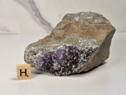 A raw amethyst geode specimen with a brown and gray crystalline outer matrix and a purple amethyst cavity, shown next to a small wooden letter tile displaying 'H4' on a light surface.
