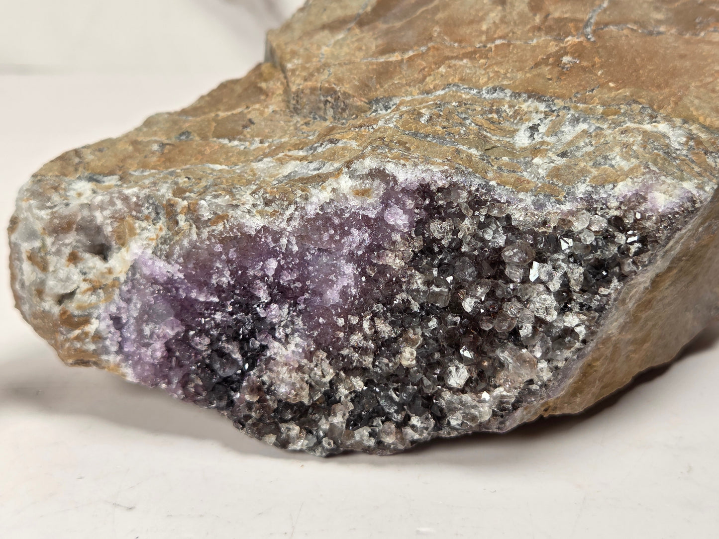 A raw mineral specimen featuring a purple amethyst crystal cluster embedded in a brown and gray crystalline rock matrix, photographed on a neutral light background.
