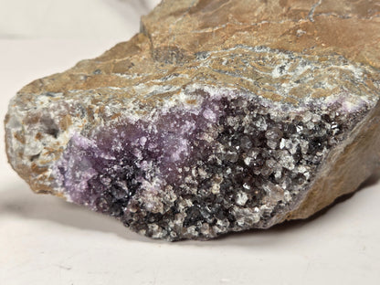 A raw mineral specimen featuring a purple amethyst crystal cluster embedded in a brown and gray crystalline rock matrix, photographed on a neutral light background.
