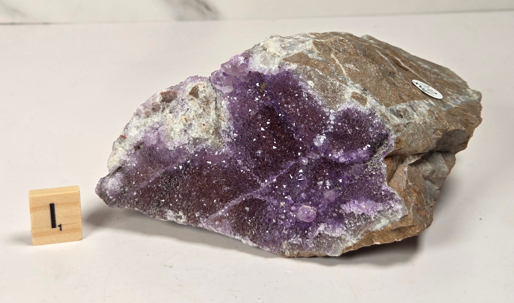 A raw amethyst geode specimen with a purple crystal cavity and white matrix, displayed on a light surface next to a small wooden block marker.