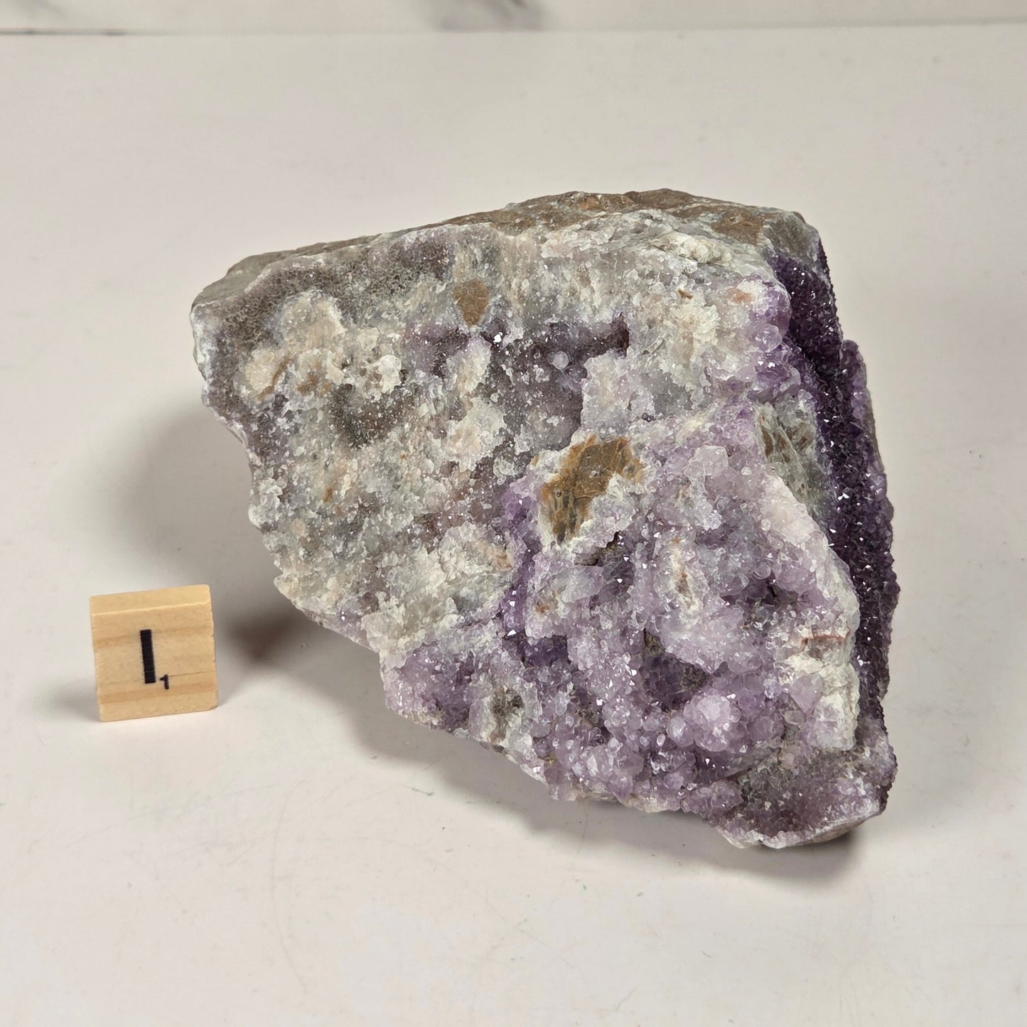 A raw amethyst crystal cluster with a rough, natural rough-hewn surface, displayed on a light gray surface next to a small wooden block scale marker.