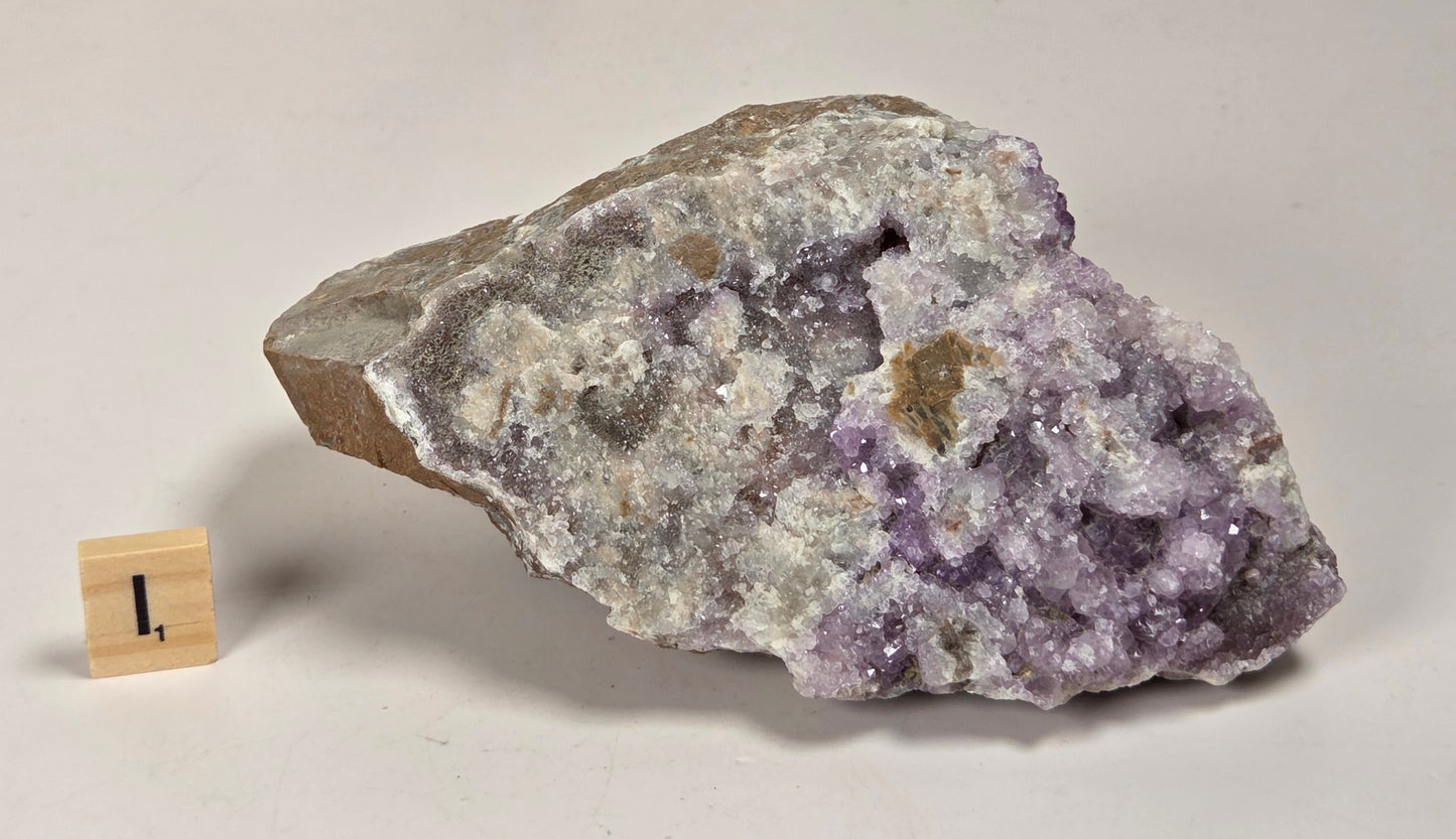 A raw amethyst geode specimen with purple crystal clusters and a brown matrix, displayed on a white surface next to a small wooden block with the number 1.