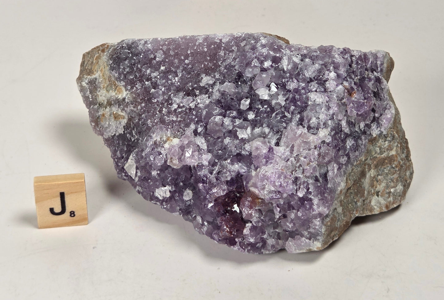 A raw amethyst geode specimen with purple crystalline clusters and gray-brown matrix, displayed on a white surface next to a small wooden letter block spelling 'Ja'.