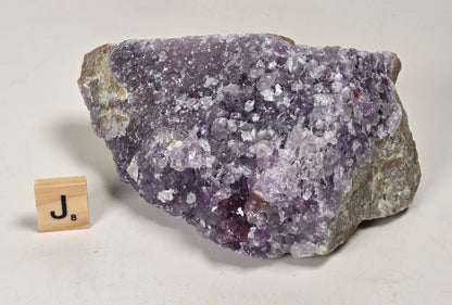 A raw amethyst geode specimen with purple crystalline clusters and gray-brown matrix, displayed on a white surface next to a small wooden letter block spelling 'Ja'.