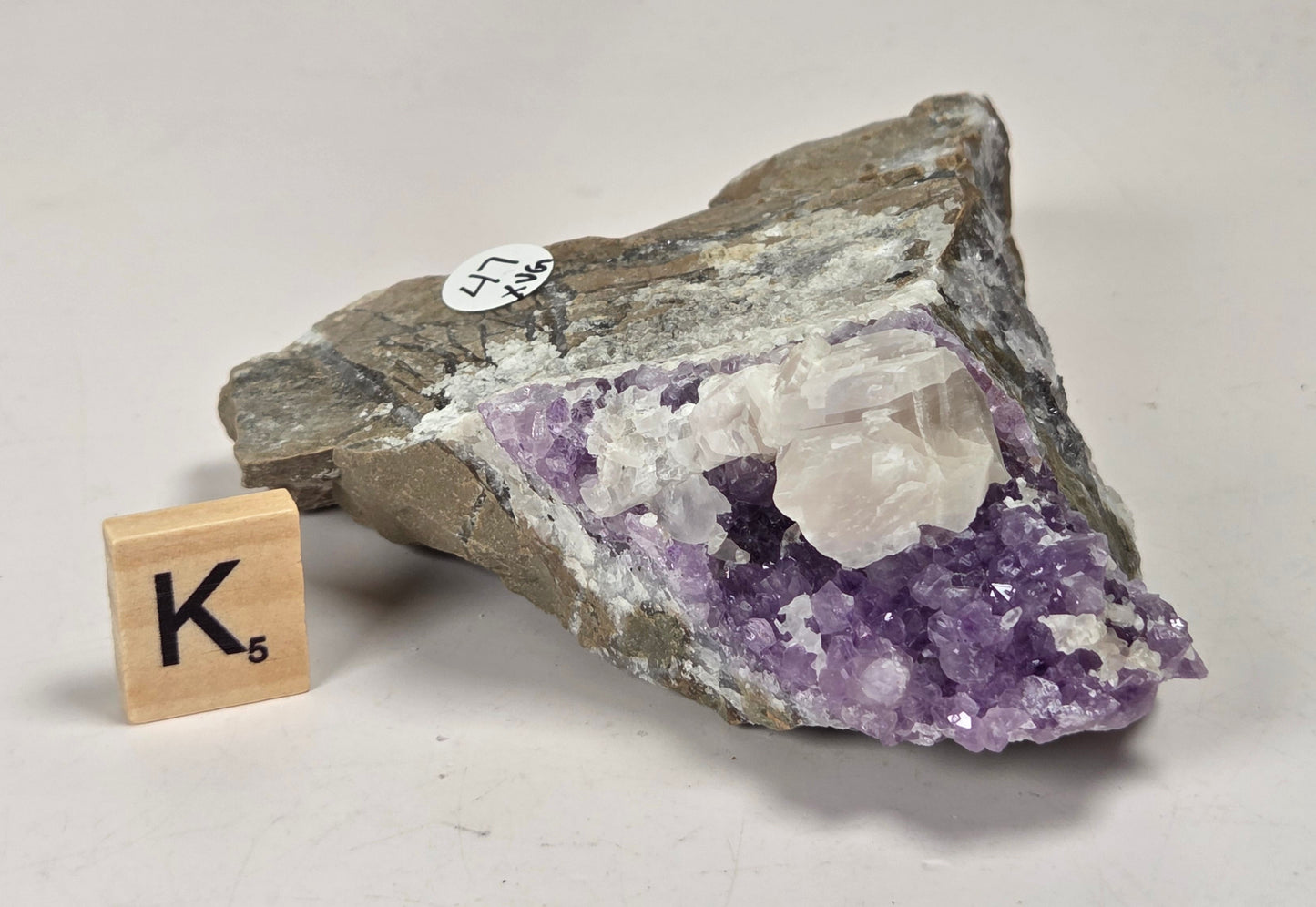 A raw amethyst crystal geode specimen with purple amethyst clusters and white quartz crystals on a rough gray-brown matrix, displayed on a light surface next to a small wooden letter tile.