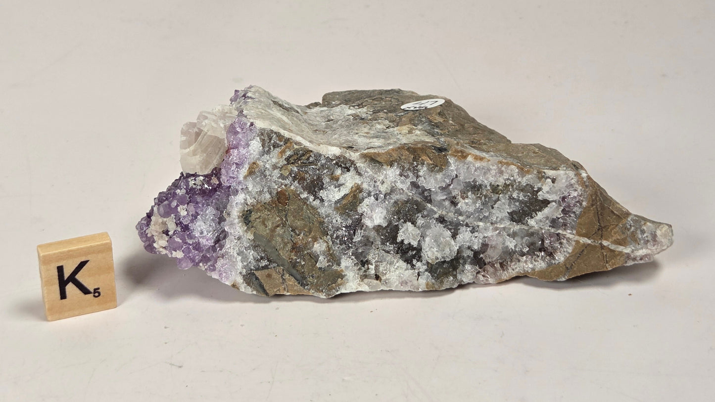A raw amethyst crystal cluster with purple and white quartz formations and gray-brown matrix, displayed on a light surface next to a small wooden Scrabble tile.