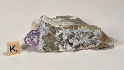A raw amethyst crystal cluster with purple and white quartz formations and gray-brown matrix, displayed on a light surface next to a small wooden Scrabble tile.