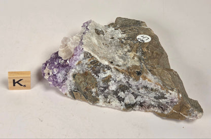 A raw mineral specimen showing a purple amethyst crystal cluster on a gray and brown matrix, displayed next to a small wooden block labeled 'K' on a plain off-white background.