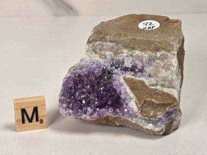 A raw amethyst geode specimen with a purple crystal cavity and brown outer matrix, displayed on a light surface next to a small wooden tile block labeled M3.