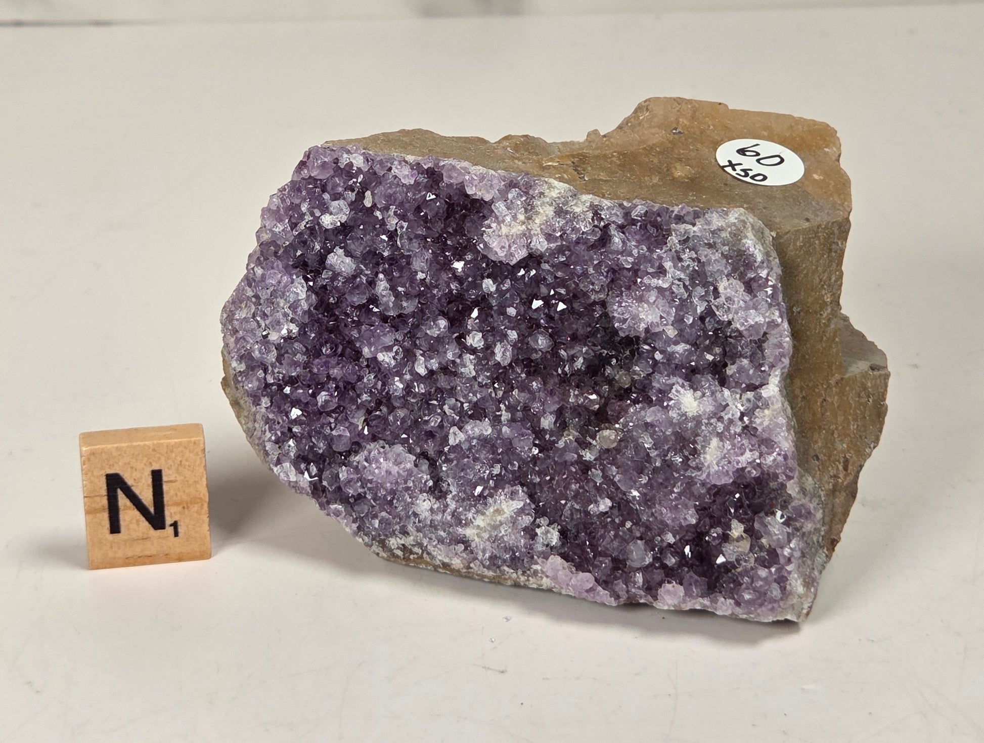 A raw amethyst geode specimen with a rough brown matrix, displayed on a white surface next to a wooden letter tile showing 'N', with a small price sticker on the rock.