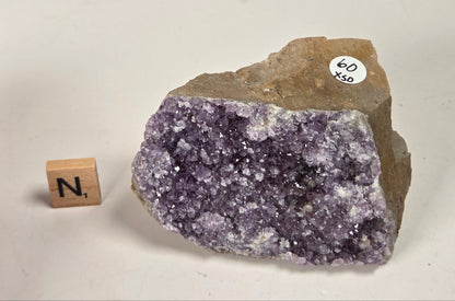 A raw amethyst crystal cluster specimen with a rough brown matrix, shown on a light beige background next to a small wooden block with the letter 'N' and a white circular sticker reading '60 x 350'.